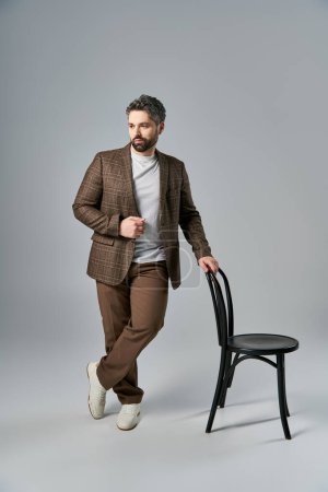 Ein stilvoller Mann mit Bart steht neben einem eleganten schwarzen Stuhl in einem Studio-Ambiente.