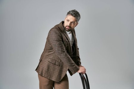 Un homme barbu suave frappant une pose tout en tenant une chaise, vêtu d'une tenue élégante sur un fond de studio gris.