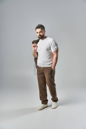 Ein stilvoller Mann mit Bart steht selbstbewusst mit der Hand in der Tasche in eleganter Kleidung vor grauem Hintergrund in einem Atelier.