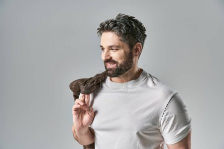 Foto de Un hombre elegante con barba sostiene una chaqueta en una pose cautivadora sobre un fondo gris de estudio. - Imagen libre de derechos