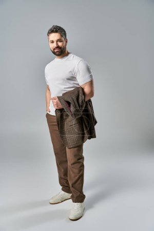 A bearded man exudes elegance in a white t-shirt and brown pants against a grey studio background.