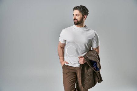 A stylish man with a beard poses confidently in elegant white t-shirt and brown pants against a grey studio background.