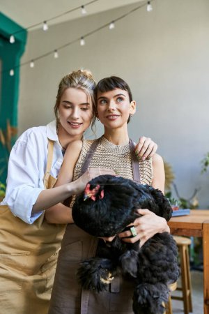 Zwei Frauen, ein liebendes lesbisches Paar, stehen zusammen in einem Kunstatelier und halten eine Henne.