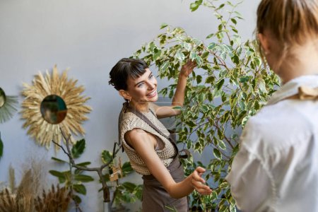 Lesbian couple in an art studio, one woman standing next to another holding a plant.
