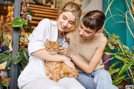Foto de Dos mujeres, una pareja lesbiana cariñosa, se sientan tranquilamente en un porche con un gato, rodeado de decoración artística. - Imagen libre de derechos