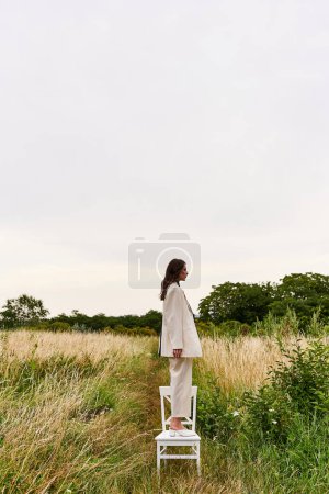 Foto de Una hermosa joven vestida de blanco se para en una silla, disfrutando de la brisa del verano en un campo. - Imagen libre de derechos