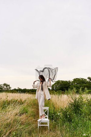 Foto de Una elegante joven vestida de blanco se para en una silla, disfrutando de la brisa del verano en un paisaje de campo. - Imagen libre de derechos