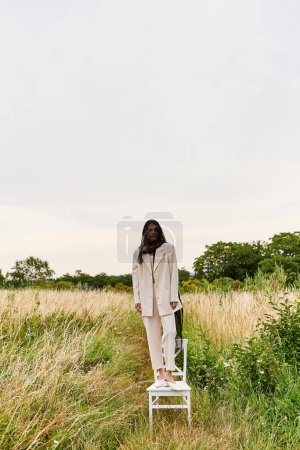 Téléchargez les photos : A beautiful young woman in white attire stands on a chair in a vibrant field, embracing the summer breeze. - en image libre de droit