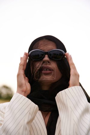 Foto de Una joven mujer exuda elegancia en un velo y gafas de sol, empapándose en la brisa del verano en un campo pintoresco. - Imagen libre de derechos