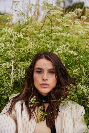 Eine schöne junge Frau in weißem Gewand steht anmutig auf einem Feld aus hohem Gras und umarmt die sanfte Sommerbrise.