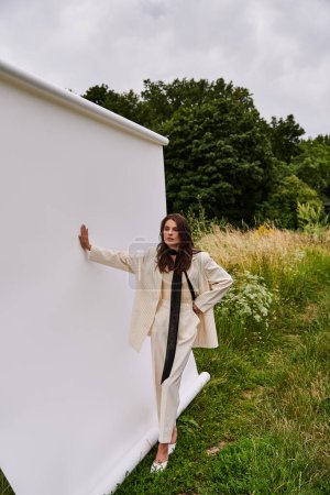Téléchargez les photos : Une jeune femme en tenue blanche se penche contre un mur blanc, embrassant la brise d'été dans un cadre de champ serein. - en image libre de droit