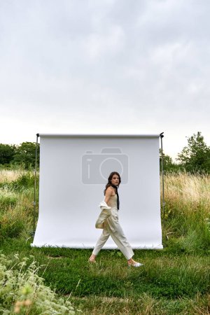A beautiful young woman in white attire gracefully walks in front of a white backdrop, enjoying the summer breeze in a field.