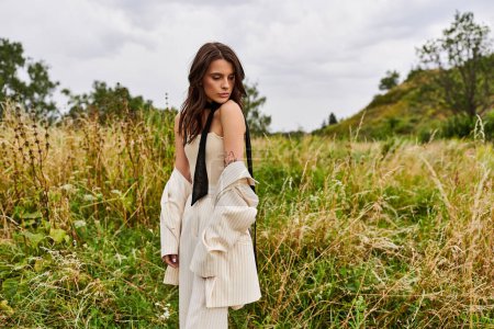 Eine schöne junge Frau in weißem Gewand steht hoch oben auf einem Feld mit hohem Gras und umarmt die sanfte Sommerbrise.