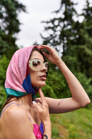 Téléchargez les photos : Une belle jeune femme dans une écharpe rose et bleue vibrante et des lunettes de soleil élégantes, profitant de la brise estivale dans la nature. - en image libre de droit