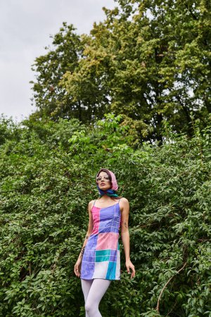 Foto de Una hermosa joven con un vestido vibrante y gafas de sol disfrutando de la brisa del verano frente a exuberantes árboles. - Imagen libre de derechos