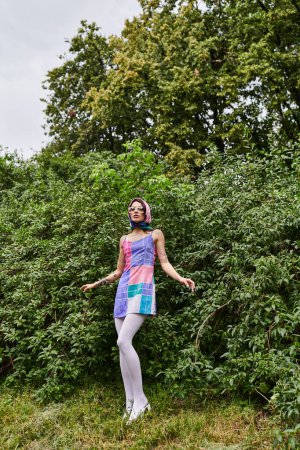 Foto de Una mujer joven en un vestido vibrante y gafas de sol se encuentra en un campo con árboles en el fondo, empapado en la brisa de verano. - Imagen libre de derechos