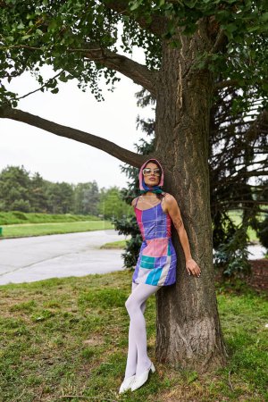Una hermosa joven con un vestido vibrante y gafas de sol se encuentra junto a un árbol, disfrutando de la brisa del verano en un parque.
