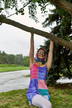 Foto de Una hermosa joven con un vestido colorido se sienta con gracia en una rama de árbol, abrazando la brisa del verano. - Imagen libre de derechos