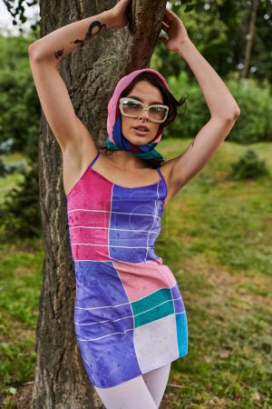Téléchargez les photos : Une jeune femme, vêtue d'une robe vibrante et de lunettes de soleil, se tient gracieusement à côté d'un arbre, immergée dans la brise estivale. - en image libre de droit