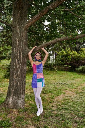 Foto de Una hermosa joven con un vestido vibrante y gafas de sol se encuentra junto a un árbol, disfrutando de la brisa del verano en un campo tranquilo. - Imagen libre de derechos