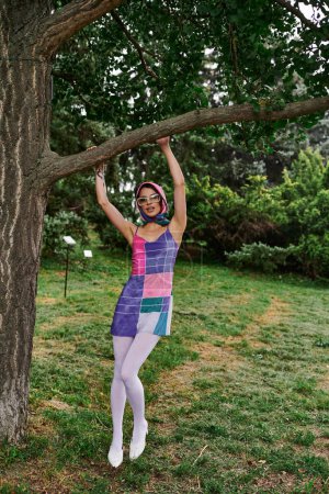 Téléchargez les photos : Une femme vibrante se tient gracieusement à côté d'un arbre majestueux dans un champ serein, se prélassant dans la brise d'été. - en image libre de droit