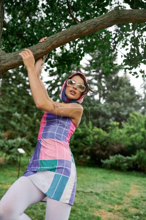 A beautiful young woman in a vibrant dress and sunglasses balancing gracefully on a tree branch, enjoying the summer breeze.