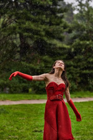 An attractive young woman in a red dress and long gloves stands gracefully in the rain, enjoying the summer downpour.
