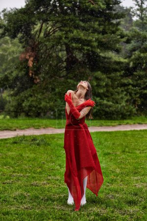 Eine bezaubernde junge Frau in einem leuchtend roten Kleid steht anmutig im Regen und umarmt die Elemente mit Gelassenheit und Eleganz.