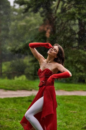 Eine junge Frau in rotem Kleid und langen Handschuhen steht anmutig im Regen und umarmt die Sommerbrise in der Natur.