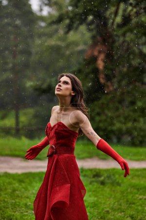 A stunning young woman in a striking red dress stands gracefully under the rain, capturing the beauty of the moment.