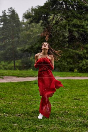 A captivating young woman in a red dress and long gloves gracefully stands in the rain, embracing the summer shower.