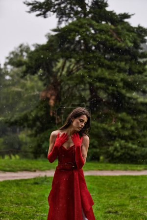 Eine atemberaubende junge Frau in rotem Kleid und langen Handschuhen steht anmutig unter einer sommerlichen Regendusche.