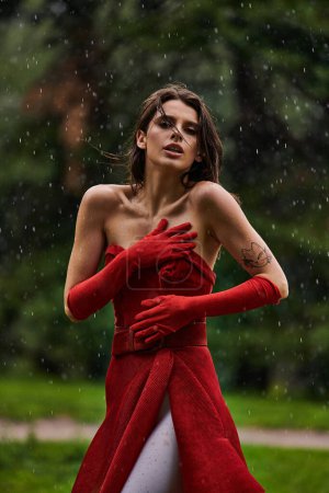 A stunning young woman in a red dress and gloves stands gracefully in the rain, embracing the elements with poise and beauty.