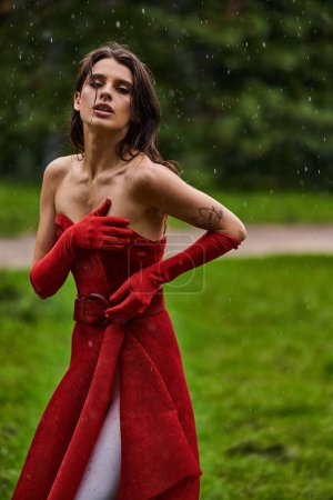 A beautiful young woman in a vibrant red dress stands gracefully in the midst of a gentle rain shower, exuding elegance.