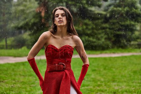 A stunning young woman in a red dress elegantly stands under the rain, embracing the moment with grace and confidence.