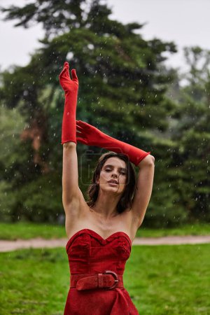 A beautiful young woman in a red dress and long gloves revels in the summer breeze in nature.