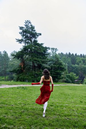 mujer joven en un vestido rojo que fluye y guantes largos corriendo alegremente a través de un campo exuberante, que encarna la libertad y el movimiento.