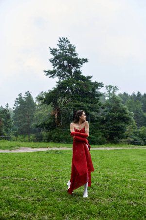 A beautiful young woman in a striking red dress and long gloves stands gracefully in a lush field, savoring the summer breeze.
