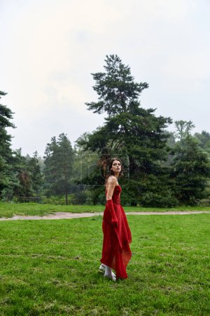 Eine attraktive junge Frau in auffallend rotem Kleid und langen Handschuhen steht anmutig in einem sonnenbeschienenen Feld und spürt die Sommerbrise.