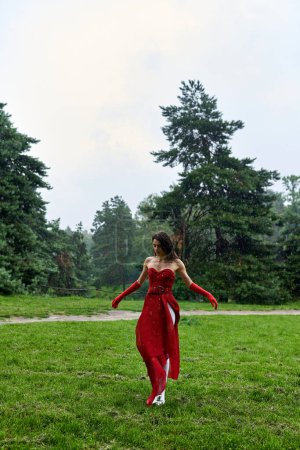 Foto de Una joven con un vestido rojo carmesí y guantes largos se levanta con gracia en un campo vibrante, abrazando la brisa del verano. - Imagen libre de derechos