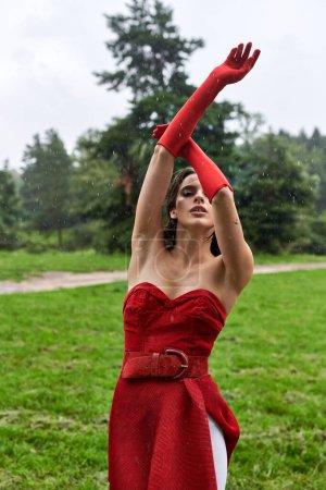 An attractive young woman in a red dress and long gloves twirls gracefully, enjoying the summer breeze in nature.