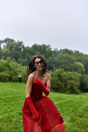 A stunning young woman stands gracefully in a field, dressed in a flowing red gown and long gloves, embracing the summer breeze.