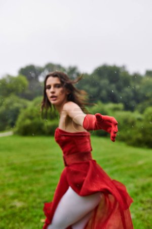 Una joven elegante con un vestido rojo vibrante y guantes largos gira alegremente en medio de la belleza natural de un campo iluminado por el sol.