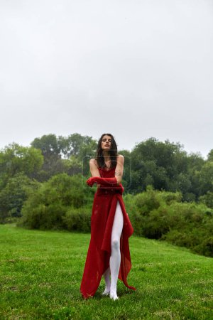 A vibrant woman in a red dress and long gloves gracefully stands in a field, immersed in the gentle summer breeze.