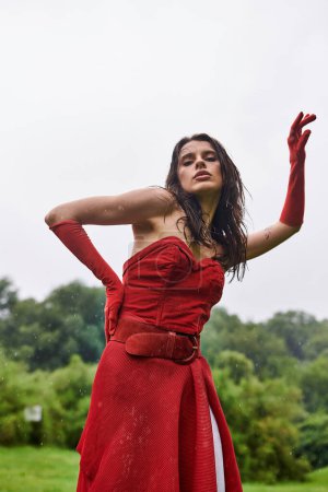 Beautiful young woman in a red dress and long gloves standing gracefully in a serene field, enjoying the summer breeze.