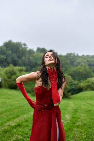 A young woman in a red dress and long gloves stands gracefully in a field, enjoying the summer breeze.