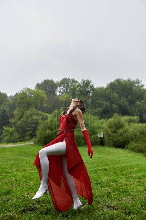 An elegant young woman in a flowing red dress and long gloves standing gracefully in a lush green field.