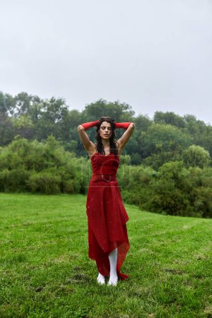 A stunning young woman in a flowing red dress and long gloves stands gracefully in a sun-kissed field, enjoying the summer breeze.