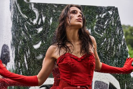 Foto de Una joven con un vestido rojo y guantes largos baila con gracia bajo la lluvia, abrazando la refrescante ducha de verano. - Imagen libre de derechos