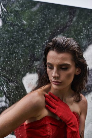 Une jeune femme en robe rouge et gants longs se tient gracieusement sous la pluie, profitant de la brise estivale dans la nature.